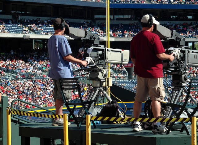 Two men experiencing fan interactions with a television set at a baseball game.